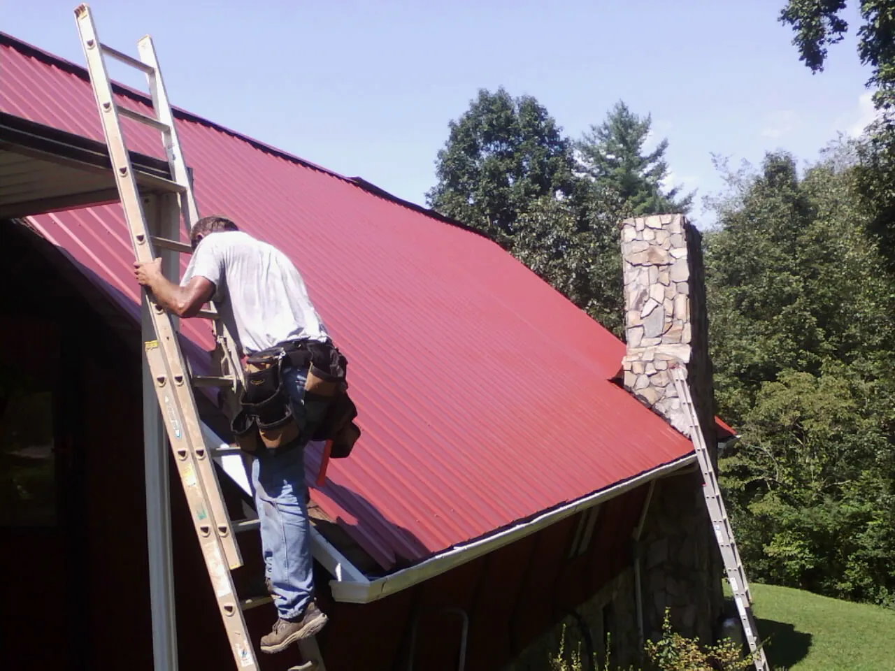 Roofer on a ladder beside a red metal roof and stone chimney — Soffit & Fascia Repair by Roofing Brothers in Shartlesville
